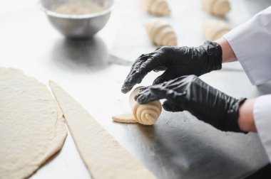 Fırıncı siyah eldivenler giyip kruvasan hazırlıyor, hamuru şekillendiriyor profesyonel mutfakta paslanmaz çelik masa üzerinde.