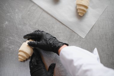 Siyah eldiven giyen fırıncı paslanmaz çelik bir masada kruvasan hamuru ustalıkla şekillendiriyor. Bu da hamur işlerinde uzman olduğunu gösteriyor.