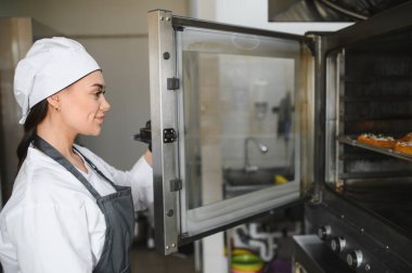 Üniformalı ve önlüklü profesyonel fırıncı büyük fırını açıyor ve ticari mutfakta hamur işlerinin ilerlemesini kontrol ediyor.