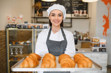 Gülümseyen fırıncı kadın büyüleyici fırınında içi yeni pişmiş, altın kruvasanlarla dolu bir tepsi sunuyor, sıcaklık ve keyif saçıyor.