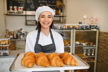 Gülümseyen fırıncı kadın elinde taze pişmiş kruvasan tepsisiyle, lezzetli kreasyonlarını bir pastanede sergiliyor.