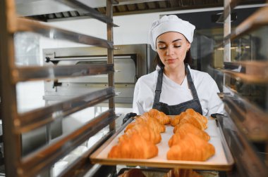 Kadın fırıncı dikkatlice altın rengi kruvasan tepsisini profesyonel bir mutfakta soğutma rafına yerleştiriyor.