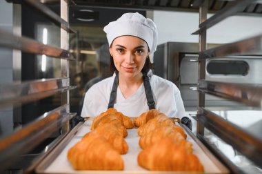 Siyah eldiven ve beyaz üniforma giyen dişi fırıncı profesyonel fırından bir tepsi altın rengi kruvasan alıyor.