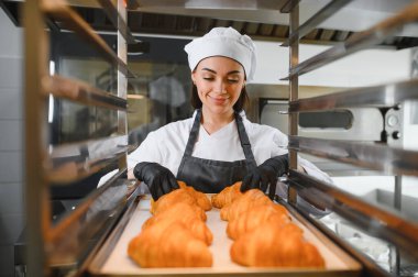 Siyah eldiven giyen dişi fırıncı profesyonel bir fırına bir tepsi altın rengi kruvasan koyuyor.