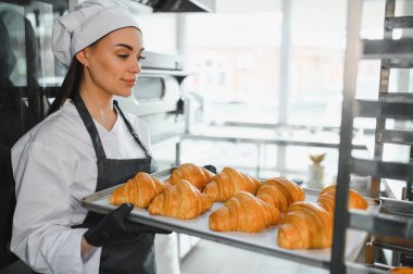 Profesyonel bir fırıncı, elinde bir tepsi altın rengi kruvasan taşıyor ve ticari bir mutfakta yaptığı pişirme uzmanlığının lezzetli sonuçlarını sergiliyor.
