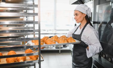 Profesyonel bir fırıncı ticari bir mutfakta elinde bir tepsi altın rengi kruvasan taşır ve pişirme uzmanlığının leziz sonuçlarını gözler önüne serer.