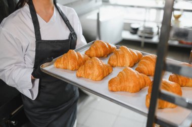 Profesyonel bir fırıncı bir tepsi altın kruvasan sunuyor. Ticari bir mutfakta titizlikle pişirilen kruvasanın sonucunu sergiliyor.
