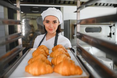 Profesyonel bir fırıncı bir tepsi altın rengi kruvasan sunuyor. Ticari bir mutfak ortamında pişirme uzmanlığının sonucunu sergiliyor.