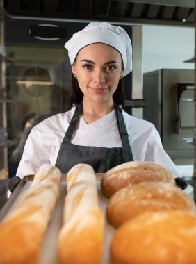 Profesyonel fırıncı fırın mutfağında içi taze pişmiş ekmek ve altın sarısı baget dolu bir tepsi sunar.