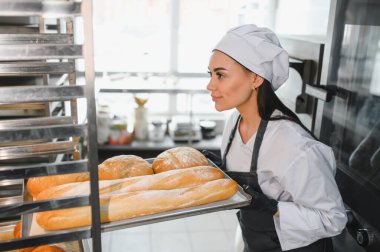 Profesyonel bir fırıncı taze pişmiş ekmek tepsisi ve yuvarlak somunları taşır ve onları ticari bir mutfaktaki metal soğutma rafına koyar.