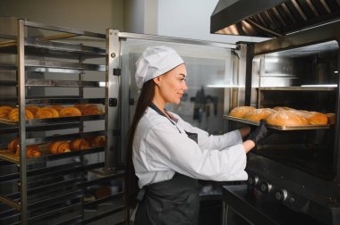 Profesyonel fırıncı, altın kahverengi somun tepsisini endüstriyel fırına yerleştiriyor. Etrafı hamur işleriyle çevrili.