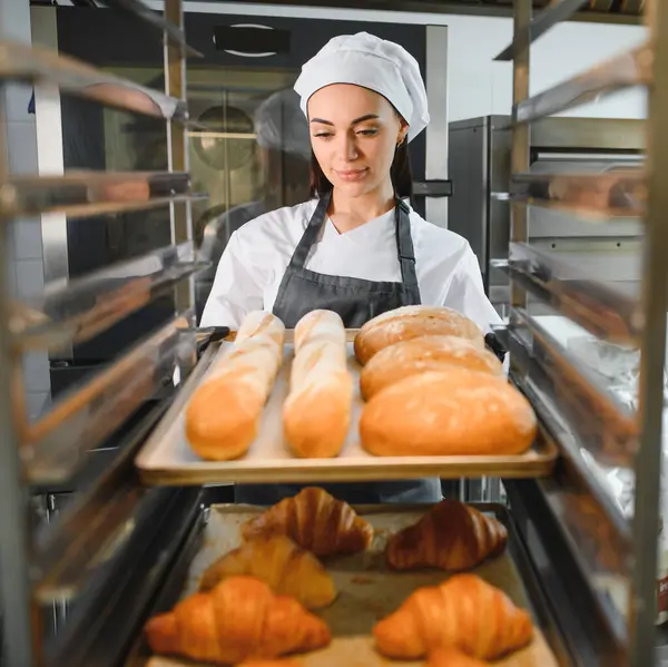 Profesyonel bir fırıncı bir tepsi baget ve yuvarlak somunları bir fırın mutfağındaki rafa yerleştiriyor ve alt rafta kruvasanlar var.