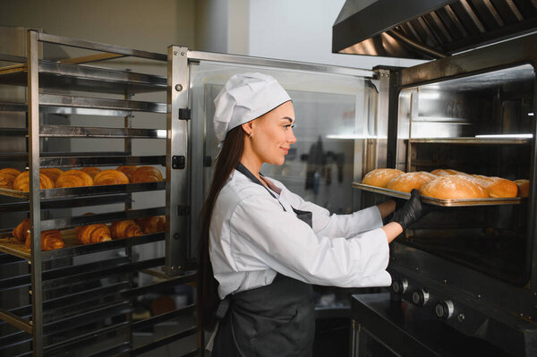 Профессиональный пекарь кладет поднос из золотисто-коричневых хлебов в промышленную печь, окруженный стойками выпечки
