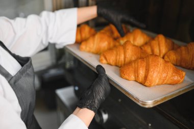 Fırıncı siyah eldiven giyip profesyonel bir fırından taze pişmiş bir tepsi altın kruvasan alıyor.