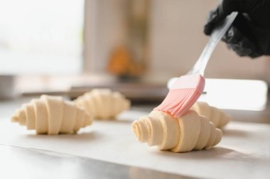 Fırıncı kruvasanları yumurta yıkama ile parlatıyor silikon fırça kullanıyor, profesyonel mutfakta pişirmeye hazırlanıyor.