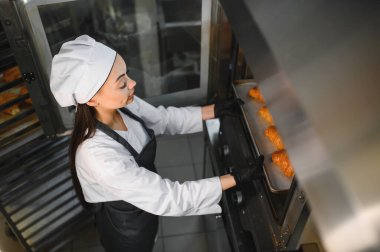 Profesyonel fırıncı siyah eldiven giyer. Fırındaki endüstriyel fırına taze kruvasan koyar.