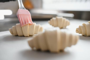 Fırıncı kruvasanları yumurta banyosuyla parlatıyor silikon fırça kullanıyor, profesyonel mutfakta pişirmeye hazırlanıyor.