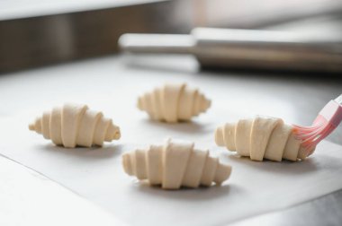 Fırıncının kruvasanların üzerinde yumurta fırçalamasına yakın çekim, profesyonel mutfakta pişirmek için hamur işleri hazırlama