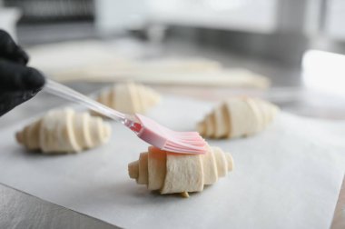 Fırıncı, taze pişmiş kruvasanları canlı pembe kremayla donatıyor. Profesyonel mutfak ortamında silikon fırça kullanıyor.