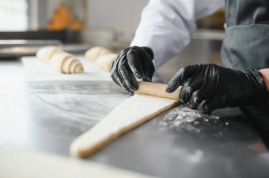 Siyah eldivenli fırıncı hamuru mükemmel bir silindir şeklinde yuvarlıyor, profesyonel bir mutfakta lezzetli kruvasanlar yapmaya hazırlanıyor.