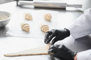 Fırın mutfağında kruvasan hamuru saran siyah eldivenli fırıncı arka planda yeni pişmiş kruvasan var.