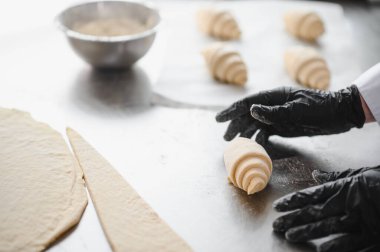 Siyah eldiven giyen fırıncı kruvasanları ustalıkla hazırlıyor, hamur şekillerini metal bir masada yuvarlıyor ve şekillendiriyor.