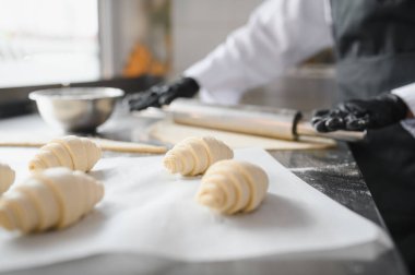 Siyah eldiven giyen fırıncı hamuru yuvarlıyor ve profesyonel mutfakta pişmemiş kruvasan hazırlıyor.