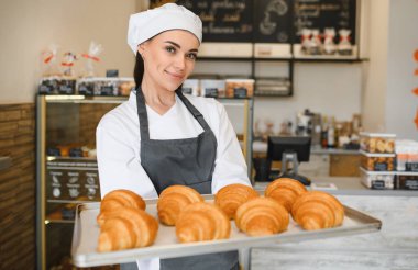 Bir tepsi lezzetli altın kruvasan sunan kadın fırıncı, bir fırın ortamında pişirme uzmanlığının sonucunu sergiliyor.