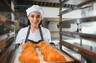 Profesyonel fırıncı gülümseyerek büyük bir sanayi fırınından yeni pişmiş altın kruvasan alıyor.