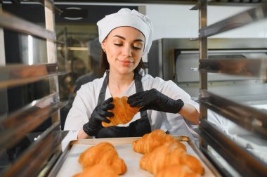 Profesyonel fırıncı siyah eldiven takıyor. Ticari bir mutfakta altın bir kruvasanı dikkatle inceliyor.