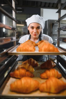 Profesyonel bir fırıncı, bir tepsi taze pişmiş altın kruvasanı ticari bir mutfakta fırından çıkarıyor.