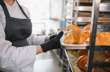 Siyah eldivenler takan fırıncı, fırının mutfağındaki metal raflara altın rengi kruvasanları dikkatlice yerleştiriyor.