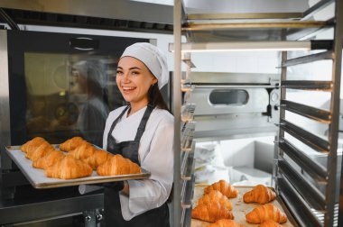 Gülümseyen fırıncı, altın rengi kruvasan tepsisini sunar. Profesyonel bir fırın ortamında yaptığı çalışmaların sonucunu gözler önüne serer.