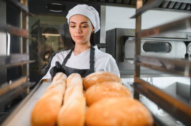 Fırıncı siyah eldiven ve önlük takıyor taze pişmiş baget ve yuvarlak somunları ticari bir fırının soğutma rafına yerleştiriyor.