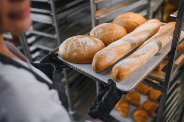 Baker dikkatli bir şekilde fırında pişirilmiş taze ekmek ve baget tepsisini rafa yerleştiriyor, fırında zanaatkar pişirme işlemi sergiliyor