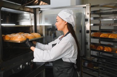 Profesyonel fırıncı, altın kahverengi somun tepsisini endüstriyel fırına yerleştiriyor. Etrafı hamur işleriyle çevrili.