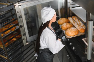 Siyah eldiven giyen profesyonel fırıncı büyük bir fırından bir tepsi taze ekmek alıyor.