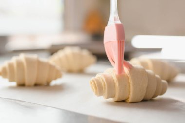 Silikon fırçası kruvasana yumurta banyosu uyguluyor, profesyonel fırında altın ve lapa kabuğu oluşturuyor.