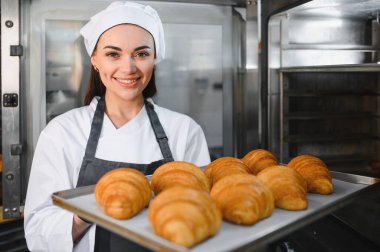Kadın fırıncı gülümsüyor ve elinde bir tepsi altın rengi kruvasan tutuyordu. Profesyonel bir mutfakta fırından yeni çıkmıştı.