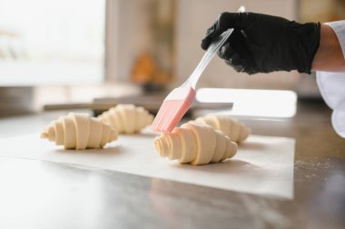 Fırıncı silikon fırçayla taze kruvasanlara yumurta banyosu yapıyor, profesyonel mutfakta pişirmek için hamur işleri hazırlıyor.