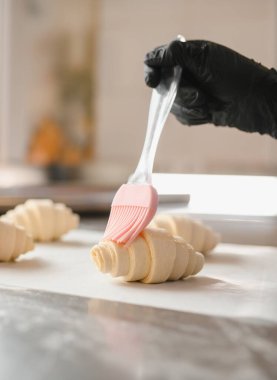 Siyah eldiven giyen fırıncı, kruvasan hamurunu pembe silikon bir fırçayla fırçalıyor ve profesyonel bir mutfakta hamur işleri hazırlıyor.