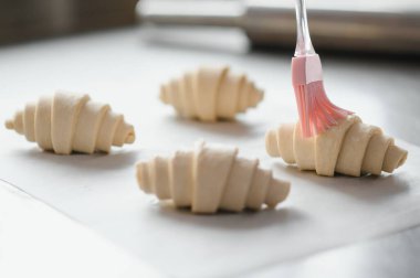 Fırıncının çiğ kruvasanlara yumurta fırçalamasına yakın çekim, profesyonel mutfakta hamur işleri hazırlama.