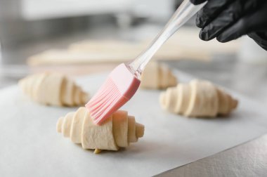 Fırıncı pişirmeden önce çiğ kruvasanlara yumurta banyosu yapıyor, profesyonel mutfakta silikon pasta fırçası kullanıyor.