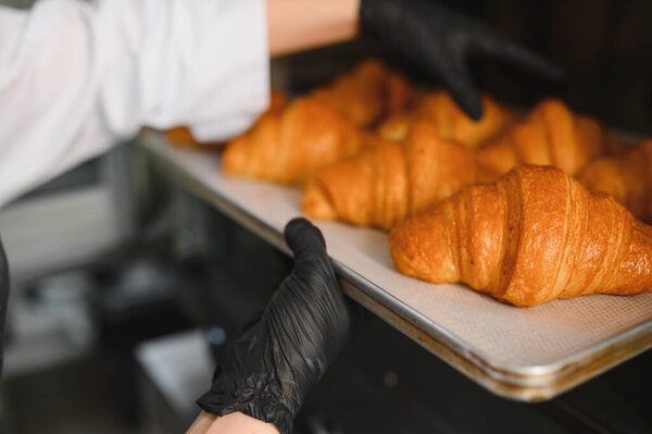 Baker wearing black gloves taking a tray of golden brown croissants out of a professional oven