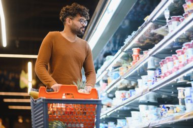Sakallı adam alışveriş arabasını itiyor ve mandıra ürünleri reyonunda yoğurt seçiyor.
