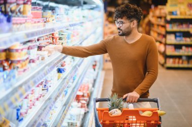 Genç Arap adam marketteki dondurulmuş raflardan yoğurt seçiyor içinde meyveler olan bir alışveriş arabasını itiyor.