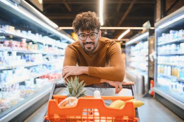 Gözlüklü genç Arap adam alışveriş arabasına yaslanıyor, gülümsüyor ve süpermarketten alışveriş yapıyor.