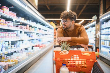 Market alışverişi yapan, alışveriş arabasına yaslanan ve dondurulmuş süt ürünlerine bakan genç bir adam.