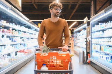 Genç Arap bir adam marketin içinde alışveriş arabasını itiyor, koridorlarda yürüyor ve diğer ürünleri arıyor.