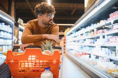 Alışveriş arabasına yaslanıp markette dondurulmuş süt ürünlerine bakan genç bir adam.